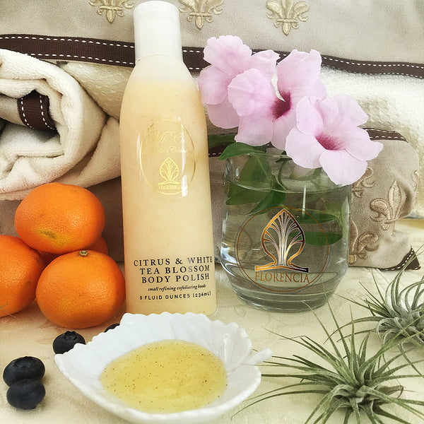 A bottle of Citrus & White Tea Blossom Body Polish resting against fabrics with oranges, blueberries and flowers next to it. Small leaf dish with the polish. 